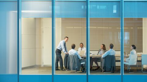 Unser Standortmanagement sichert eine wirtschaftliche und gleichzeitig attraktive Unterbringung der Landesbehörden. Außenansicht: Der Betrachter schaut durch das Fenster eines Gebäudes in einen Konferenzraum. Dort findet eine Sitzung statt – sechs Teammitglieder in Business-Kleidung tauschen sich zu einem Thema aus.