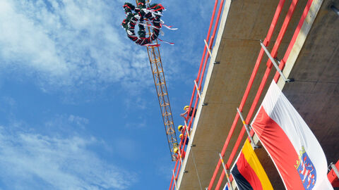 Der Landesbetrieb LBIH versteht sich als zentraler und verlässlicher Partner der Hessischen Landesverwaltung, der seine vielfältigen Baumanagement- und Immobilienbewirtschaftungs-Leistungen aus einer Hand erbringt. Auf dem Bild ist ein Teil eines Rohbaus von unten zu sehen, der einen Blick in den blauen Himmel zuläßt. An der Gebäudewand hängen Flaggen, über der Baustelle ist ein Krahn zu sehen, an dem der Richtkranz hängt.